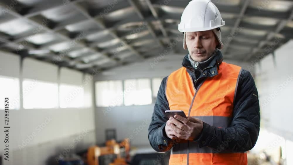man beard and mustache in a hard hat and overalls is standing in the middle of the hangar and texting message by his phone at the camera man beard and mustache in a hard hat and overalls is standing in the middle of the hangar and texting message by his phone at the camera