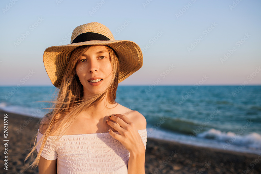Young relaxed attractive girl in straw hat spending time at the beach at sunset. Blue waves on the background.