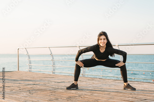 Young brunette woman with white wireless headphones in a black jumpsuit takes exercises on a pier at sunrise.