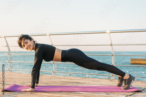 Pretty brunette young woman with white wireless headphones in a black jumpsuit takes plank workout on a mat on pier at sunrise.