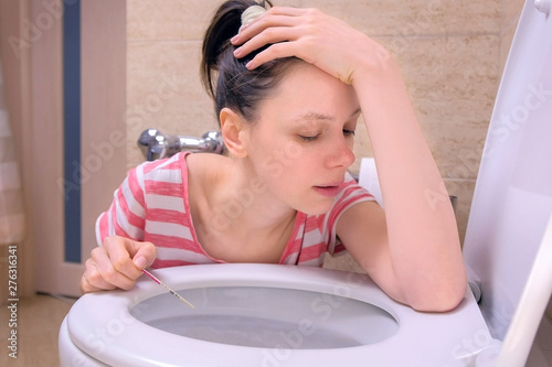 Pregnant tired woman with pregnancy test in hand is vomiting in toilet sitting on the floor at home, front view.