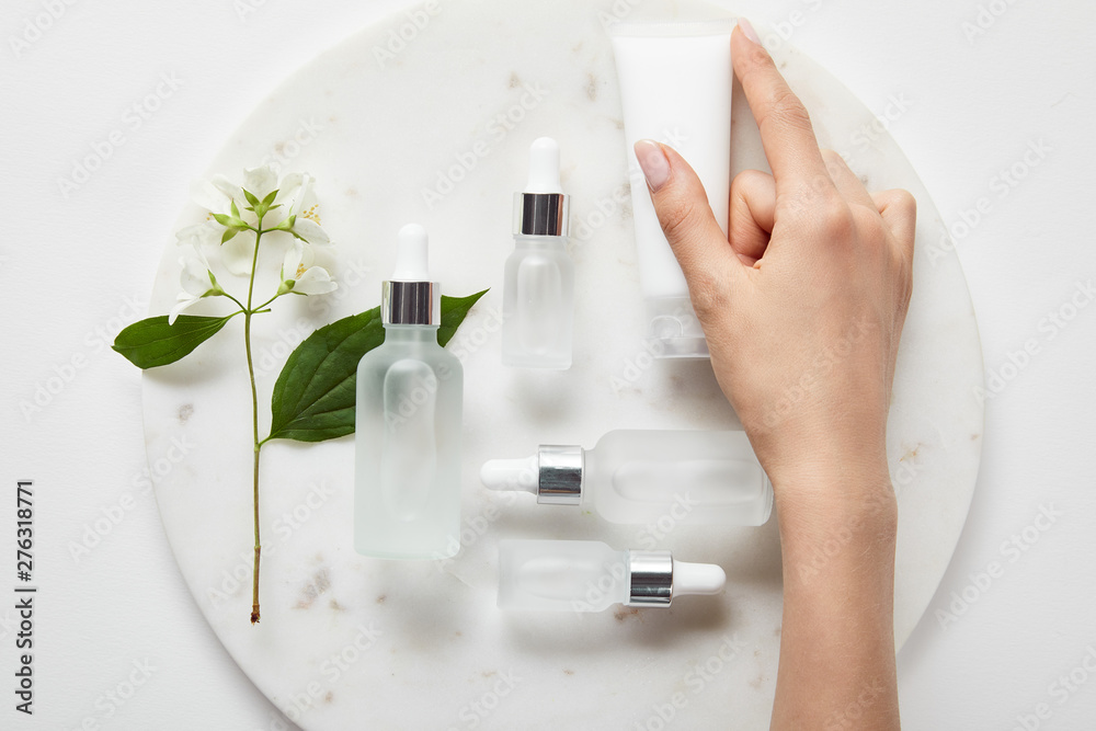 Cropped view of woman hand with cream tube over plate with jasmine and cosmetic glass bottles on white surface - obrazy, fototapety, plakaty 