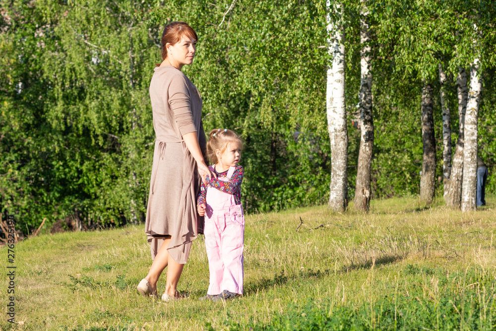 Middle aged woman holding hands with little girl in summer park with trees looking aside Middle aged woman holding hands with little girl in summer park with trees looking aside