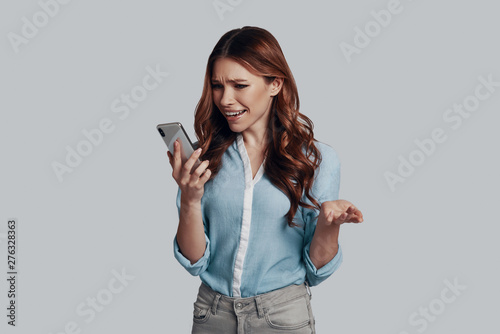 Oh my God! Surprised young woman looking at smart phone while standing against grey background