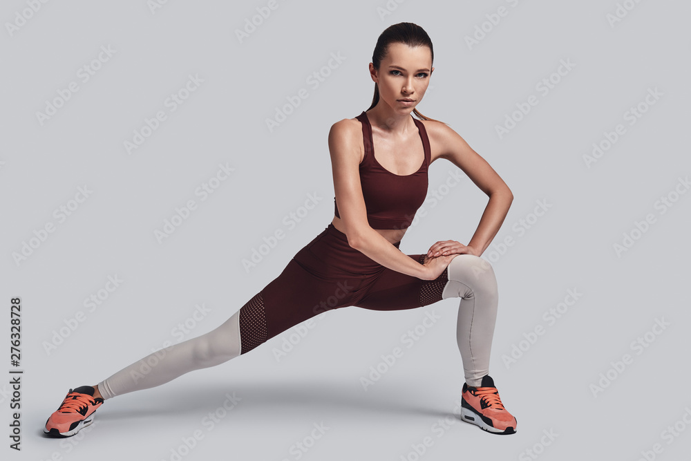 Obraz premium Full of energy. Attractive young woman stretching and looking at camera while exercising against grey background