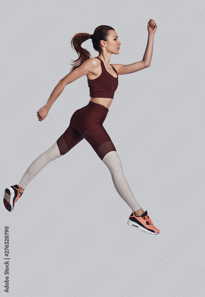 Flying high. Full length of attractive young woman in sports clothing jumping while exercising against grey background