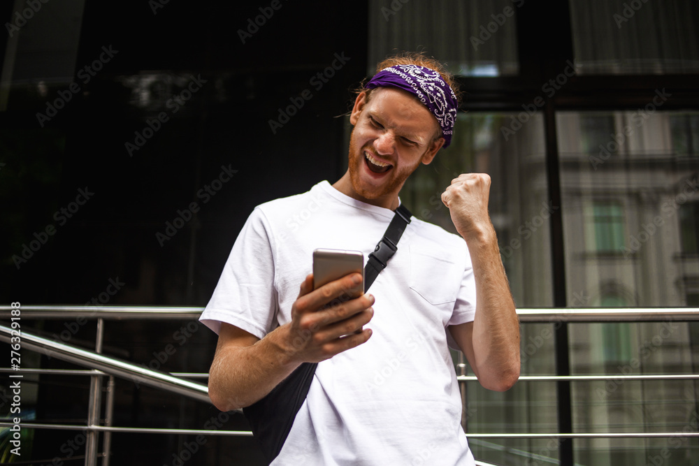 Naklejka premium Outdoor portrait of modern young man with mobile phone in the street. Excited man checking news on smart phone walking in the street. Win, winner concept. Hawing good news, wow surprised shocked face
