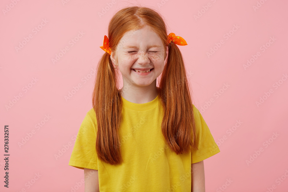 Portrait of little freckles red-haired girl with two tails, smiles with closed eyes, wears in yellow t-shirt, stands over pink background.