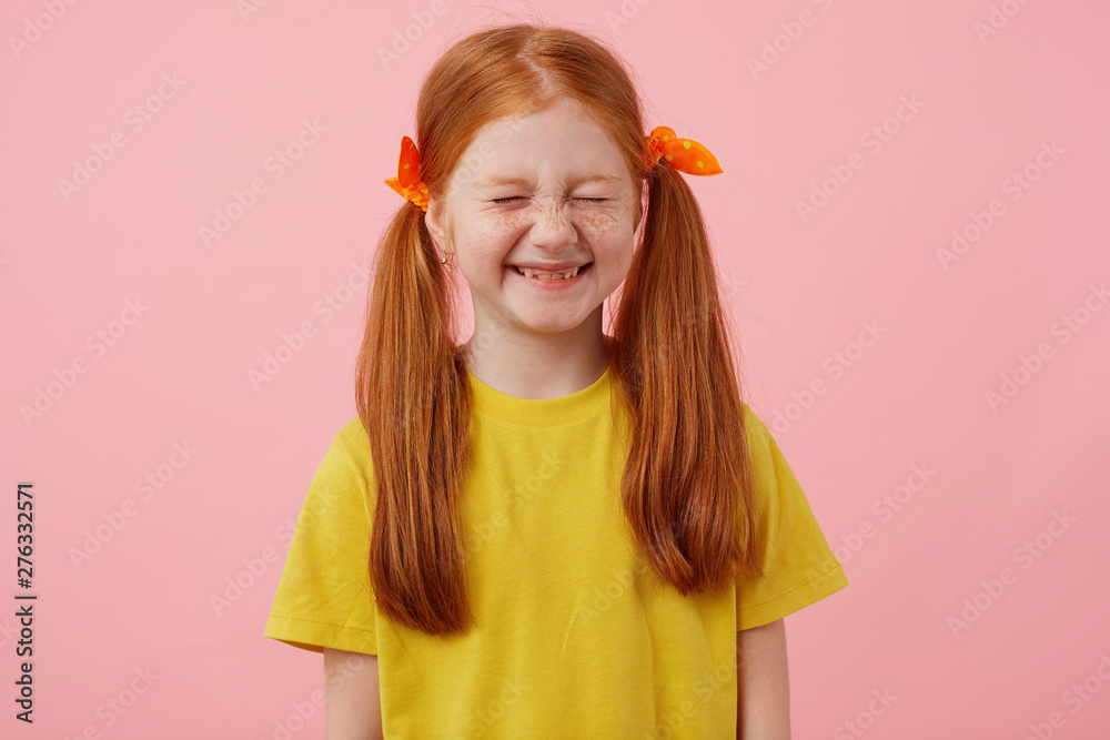 Portrait of little freckles red-haired girl with two tails, smiles with closed eyes, wears in yellow t-shirt, stands over pink background. Portrait of little freckles red-haired girl with two tails, smiles with closed eyes, wears in yellow t-shirt, stands over pink background.
