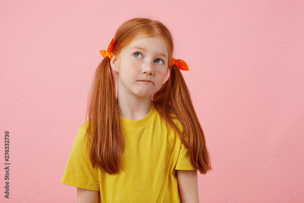 Portrait little thinking freckles red-haired girl with two tails, looks away, touches cheeks, wears in yellow t-shirt, stands over pink background with copy space. Portrait little thinking freckles red-haired girl with two tails, looks away, touches cheeks, wears in yellow t-shirt, stands over pink background with copy space.