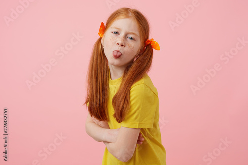 Portrait of petite freckles red-haired girl with two tails, looks and shows tounge at the camera, wears in yellow t-shirt, stands over pink background.