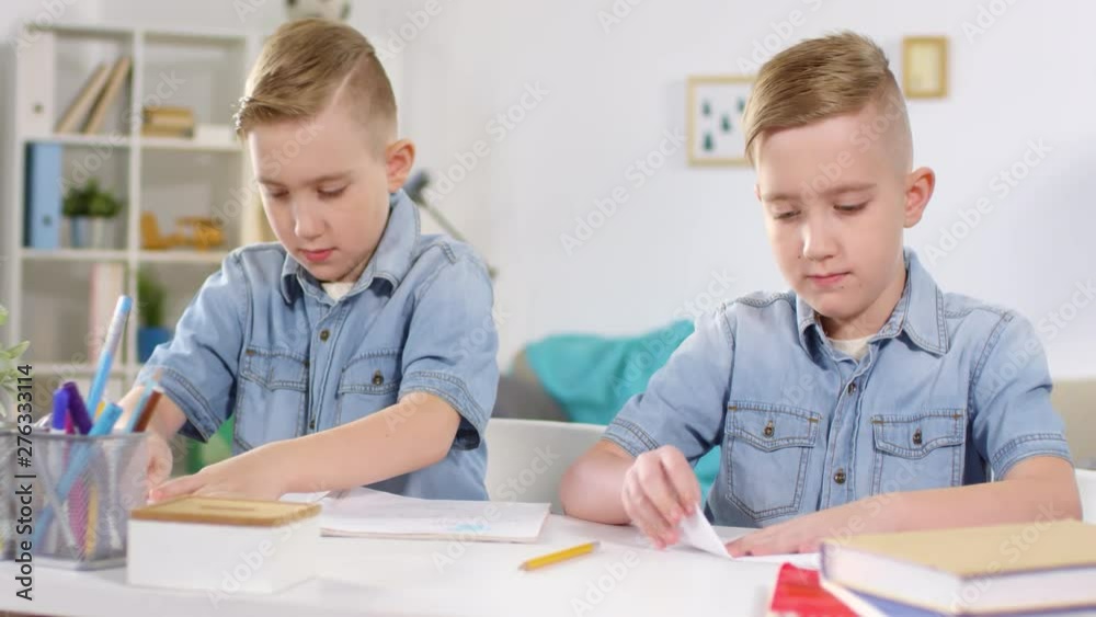 Chest-up shot of Caucasian identical twins with stylish haircuts, wearing denim shirts, sitting together at table and laboriously folding paper airplanes, throwing glances at each other’s work Chest-up shot of Caucasian identical twins with stylish haircuts, wearing denim shirts, sitting together at table and laboriously folding paper airplanes, throwing glances at each other’s work