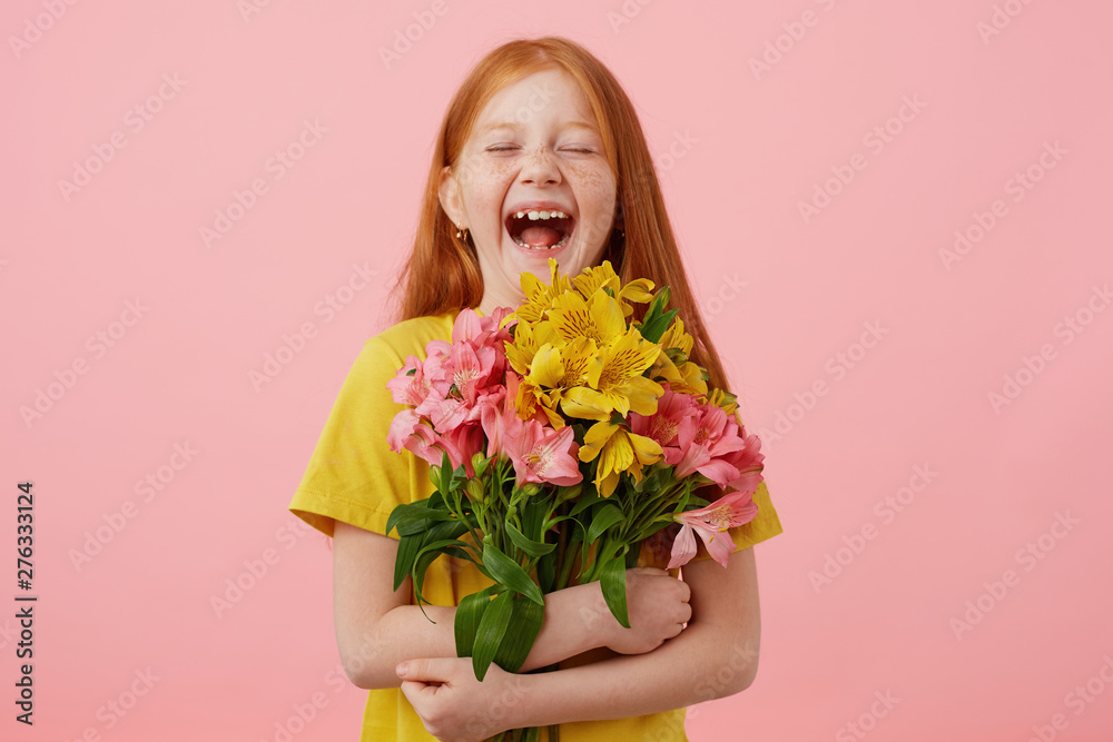 Photo of petite laughing freckles red-haired girl with two tails, with closed eyes broadly smiling and looks cute, holds bouquet, wears in yellow t-shirt, stands over pink background.