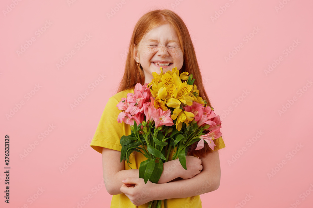 Petite freckles red-haired girl with two tails, with closed eyes broadly smiling and looks cute, holds bouquet, wears in yellow t-shirt, stands over pink background.