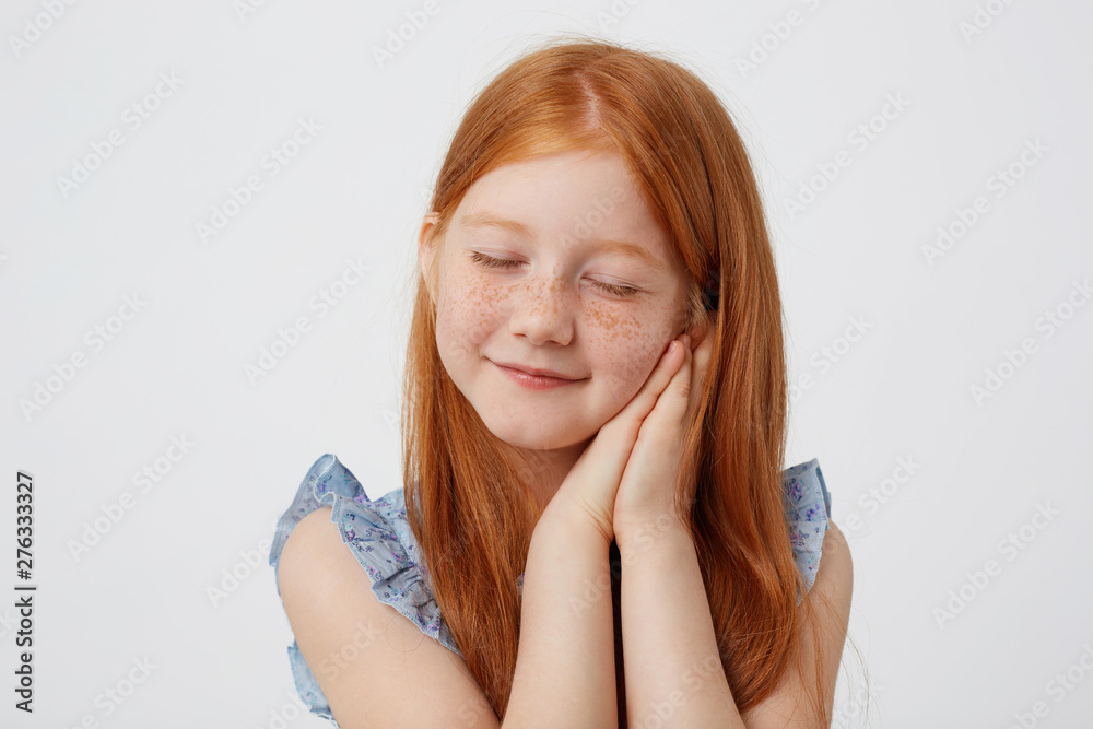 Portrait of dreaming little freckles red-haired girl in blue dress, looks sleppy, with folded palms at the cheeks, stands over white background. Portrait of dreaming little freckles red-haired girl in blue dress, looks sleppy, with folded palms at the cheeks, stands over white background.