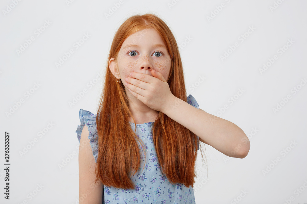 Φωτογραφία Photo of shocked little freckles red-haired, looks at the camera with wide open eyes, covers his mouth with his hand, wears in blue dress, stands over pink background