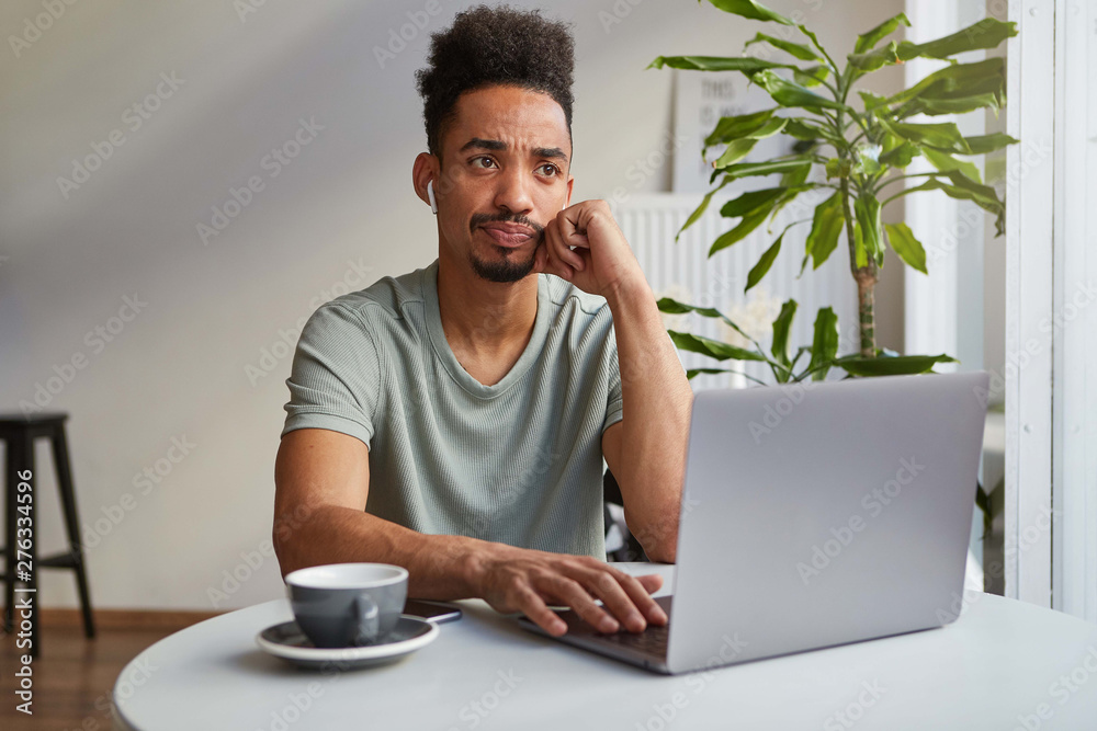 Portrait of frowning young attractive African American thinking boy, sits in a cafe, works at a laptop, touches cheek and sadly looks up, thinks about deadline.