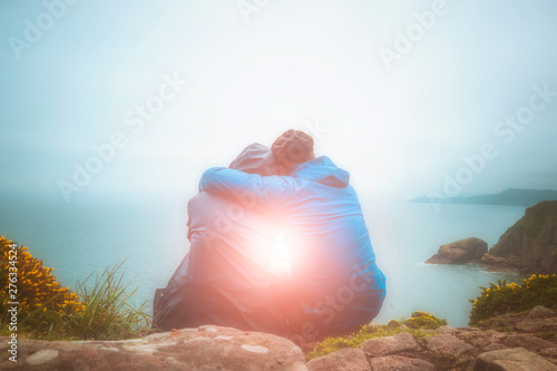 Rear or back view of young lovers sitting and hugging on a cliff edge looking at a scenic seascape with light burst. Love, romance, outdoors, nature, freedom, travel concepts.