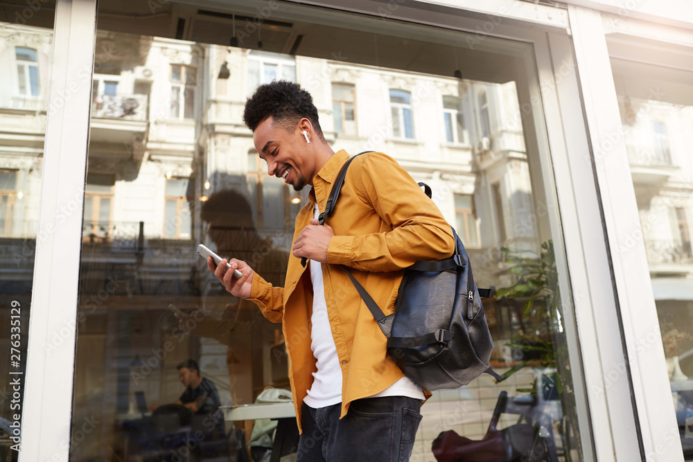 Phortrait of young cheerful dark skinned guy in yellow shirt walking down the street , holds telephone, chatting with friends, broadly smiling, looks happy. Phortrait of young cheerful dark skinned guy in yellow shirt walking down the street , holds telephone, chatting with friends, broadly smiling, looks happy.