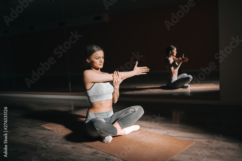 Charming sporty brunette sitting on the mat with legs crossed and stretching arm. Dark gym interior, in background mirror.