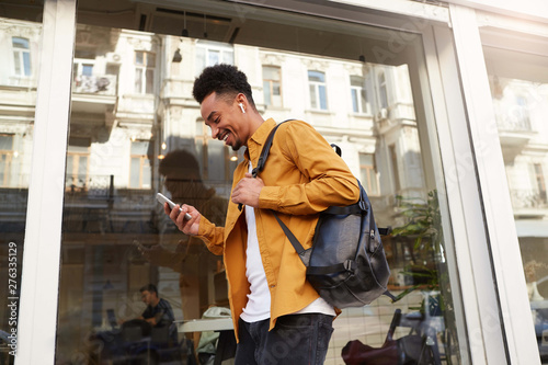 Phortrait of young cheerful dark skinned guy in yellow shirt walking down the street , holds telephone, chatting with friends, broadly smiling, looks happy.