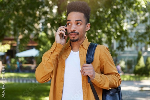 Fotografie Portrait of young calm dark skinned man in yellow shirt walking on the park , holds telephone, waiting answer from his girlfriend, looks thoughtfully