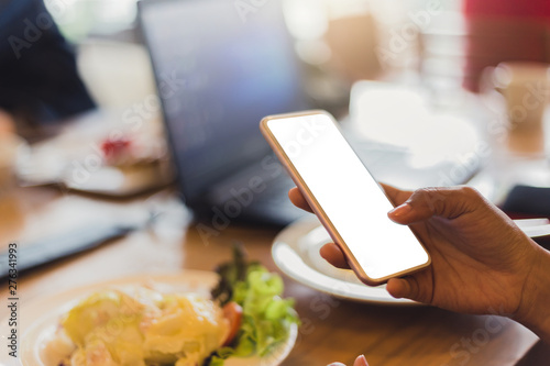 Mock up image of a women holding smartphone with blank white screen on wooden table in modern cafe restaurant during meeting or lunch.