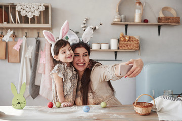 Using white colored smartphone. Mother and daughter in bunny ears at easter time have some fun in the kitchen at daytime