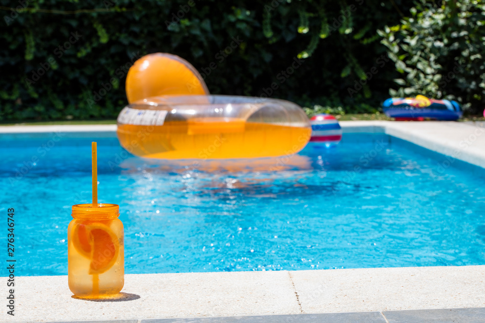 Summer cold drink. Orange mason jar on the bord of a swimming pool with big orange rubber ring floating in it