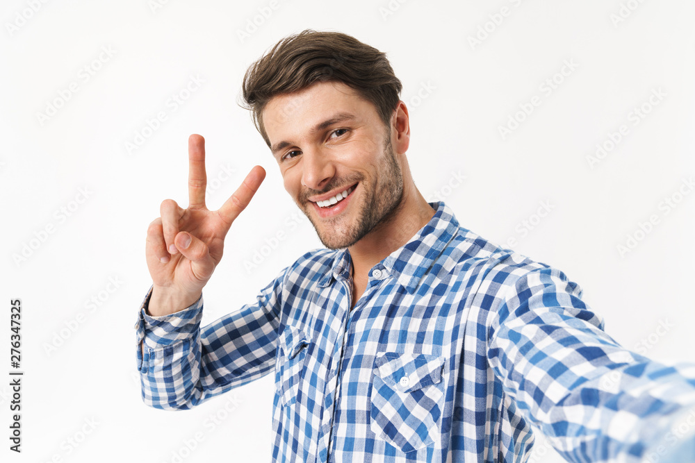 Cheerful positive bearded man dressed in casual shirt posing isolated over white wall background take a selfie by camera showing peace. Cheerful positive bearded man dressed in casual shirt posing isolated over white wall background take a selfie by camera showing peace.