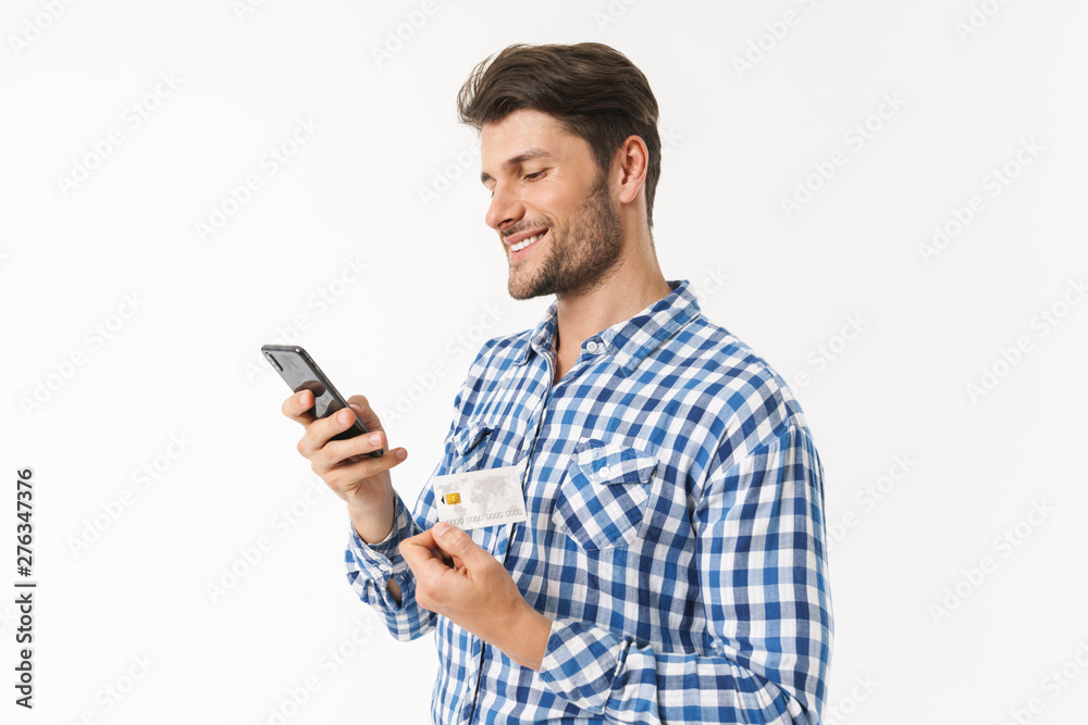 Smiling handsome young man standing isolated over white wall background dressed in casual shirt using mobile phone holding credit card. Smiling handsome young man standing isolated over white wall background dressed in casual shirt using mobile phone holding credit card.