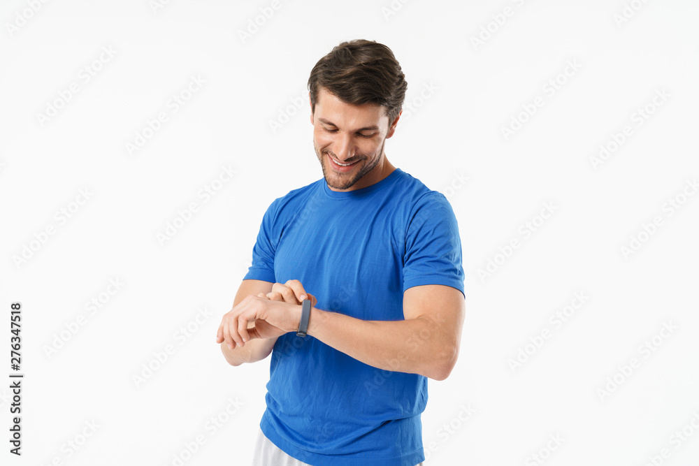 Smiling handsome young sports fitness man standing isolated over white wall background using watch clock.