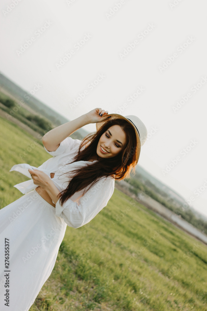 Fototapeta premium Portrait of charming carefree long hair asian girl in white clothes and straw hat enjoys life in field at sunset. Sensitivity to nature concept