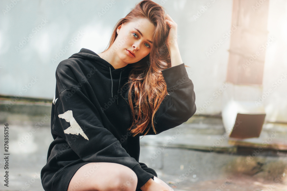 Candid portrait of young beautiful long hair unhappy girl fashion model hipster in black hoodie on wall background Candid portrait of young beautiful long hair unhappy girl fashion model hipster in black hoodie on wall background