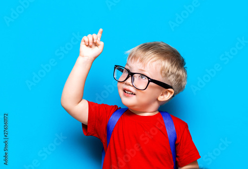 Back to school First grade junior lifestyle. Small boy in red t-shirt. Close up studio photo portrait of smiling boy in glasses with schoolbag and book show somethind with his finger