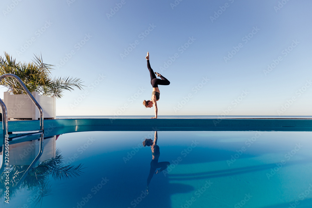 Young attractive woman practicing yoga doing Variation of handstand pose, Pincha Mayurasana. Working out by the pool, above the beach, relaxing against blue sky. Health and beauty concept