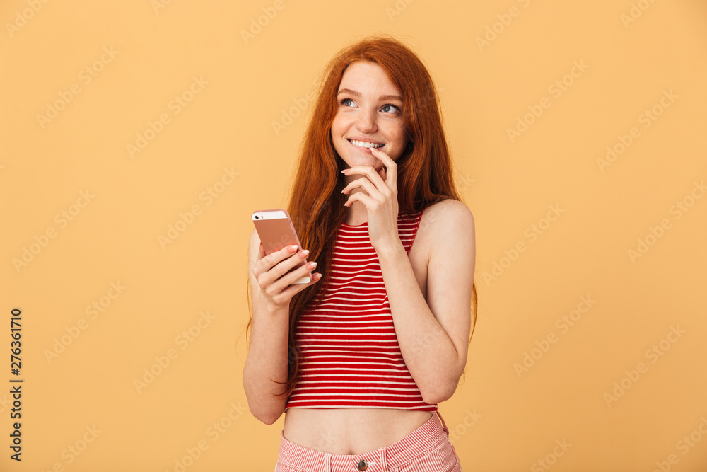 Smiling pleased cute young pretty redhead woman posing isolated over yellow background using mobile phone chatting.