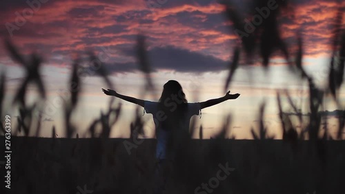 Girl with long dark hair stands alone in a field at sunset holds hands to the sides