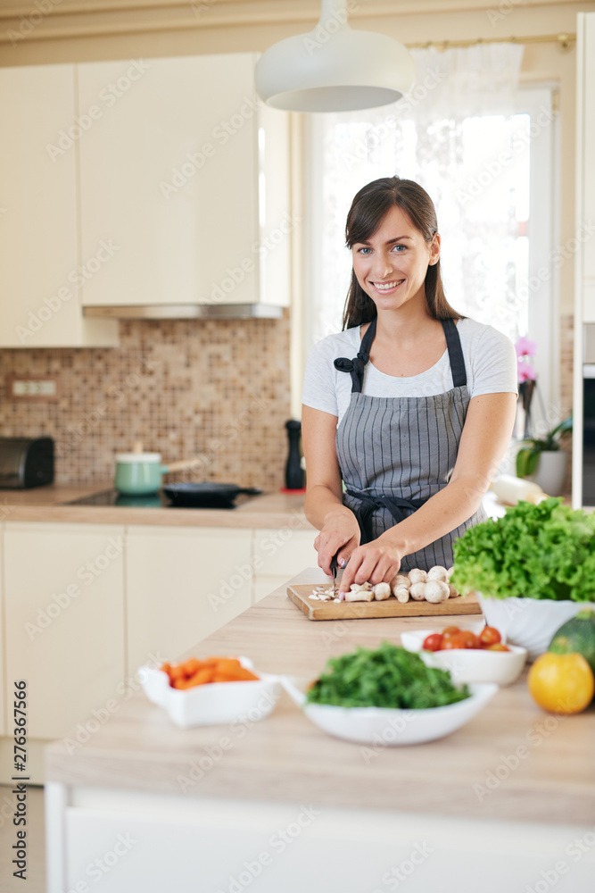 Beautiful smiling dedicated Caucasian brunette in apron standing in kitchen and chopping mushrooms. On table are lots of vegetables. Cooking at home concept. Beautiful smiling dedicated Caucasian brunette in apron standing in kitchen and chopping mushrooms. On table are lots of vegetables. Cooking at home concept.