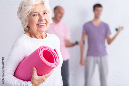 Fototapeta Naklejka Na Ścianę i Meble -  Happy senior woman holding pink joga mat, men exercising in background