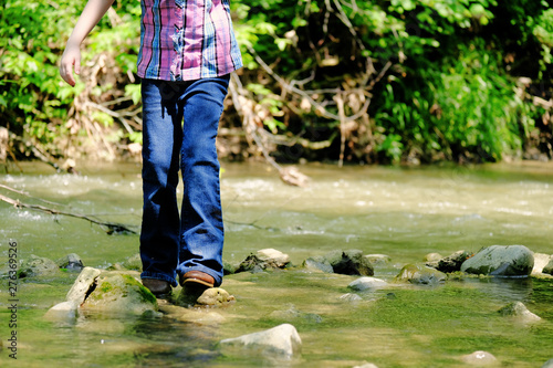 Tablou canvas Young girl in plaid shirt walking across creek outdoors with woods in background