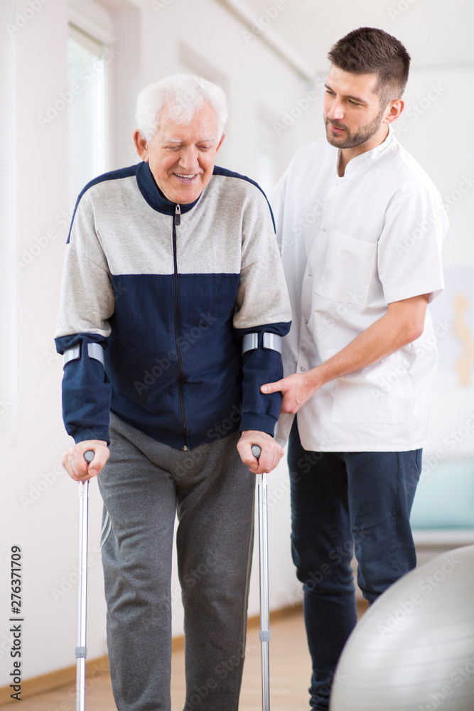 Elderly man walking on crutches and a helpful male nurse supporting him