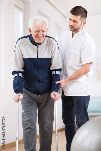 Elderly man walking on crutches and a helpful male nurse supporting him © Photographee.eu