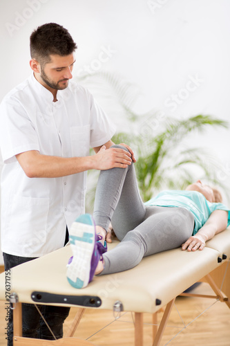 Middle age woman with knee injury lying on physiotherapy table during session with young handsome doctor