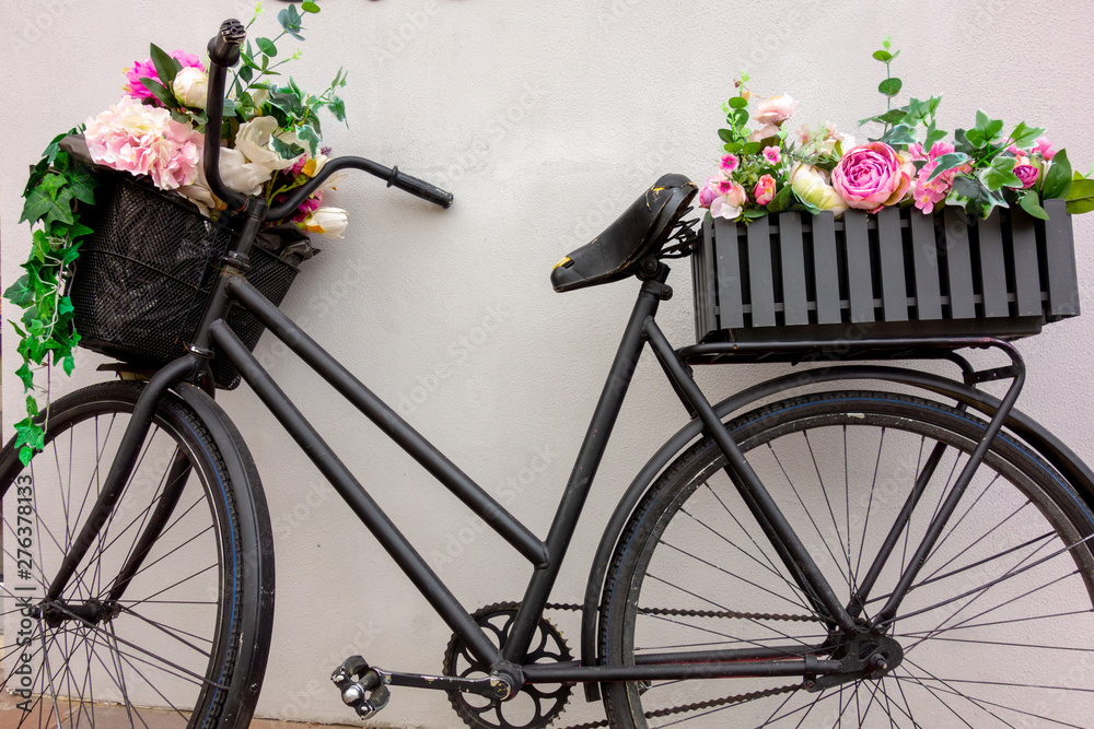 Retro bicycle as a flowerbed decoration outside element. Black aged bike stays against the wall with two garden baskets with beauty flowers. Art decor closeup