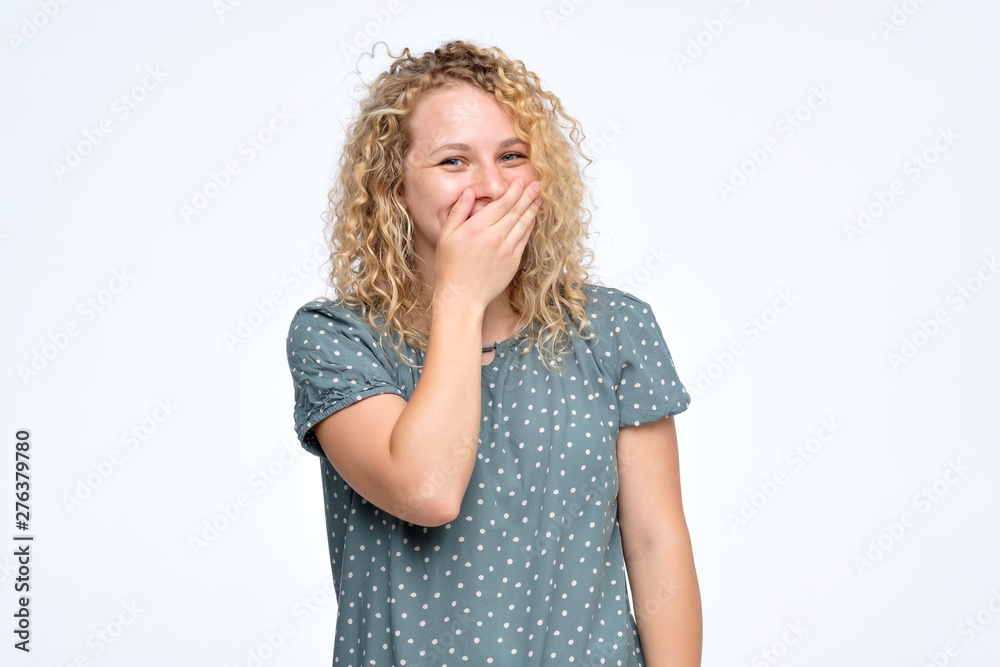 Good looking young female giggles joyfully, covers mouth as tries stop laughing. Studio shot. Positive facial emotion.