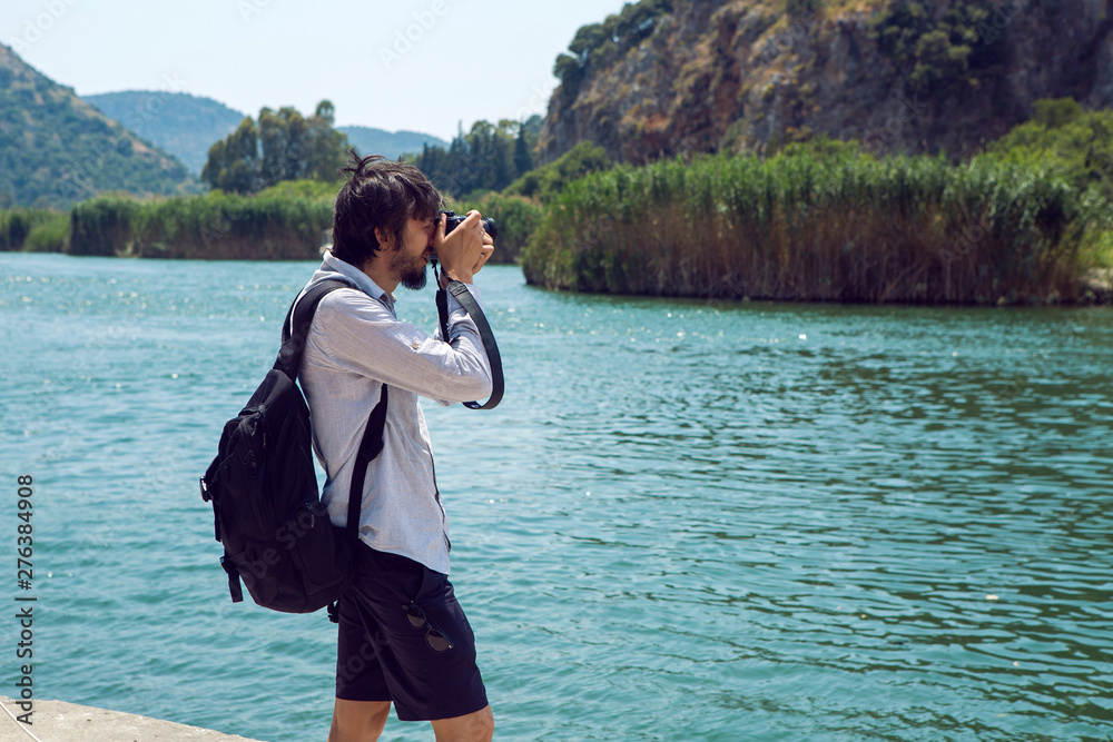 man in a shirt and shorts photographs on camera the river and the mountain