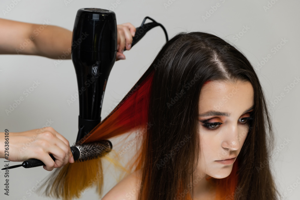 Fototapeta premium Photo portrait of the work of the master hairdresser makes a fashionable hairstyle with a hair dryer and a hairbrush in the studio on a white background. The brunette model has colored hair.