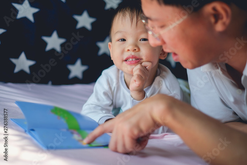 Father and Cute little Asian 2 - 3 years old toddler boy child reading bedtime story book, lying in the bed at home, Dad and son spending quality time together, Parents reading to child concept