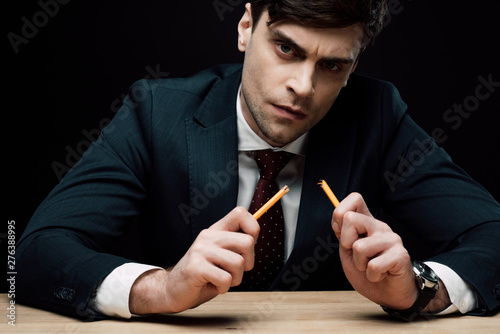 serious businessman holding broken pencil and looking at camera isolated on black photo